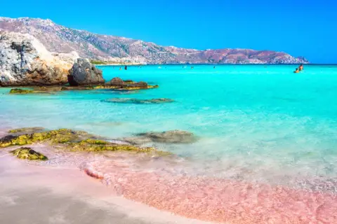 Różowa plaża Kreta z turkusową wodą i skalistym wybrzeżem. W oddali widać góry i kąpiących się ludzi.