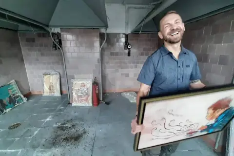 Uśmiechnięty kossakowski malarz prezentuje swoje dzieła. W tle widać obrazy i gaśnicę.
