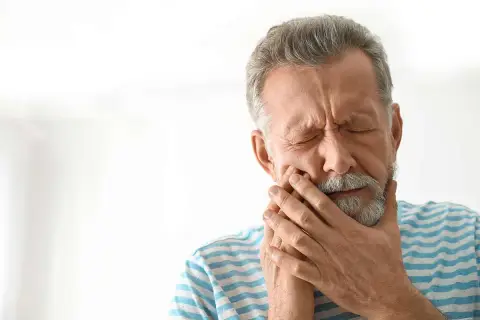 Hombre mayor con los ojos cerrados y la mano en la mejilla, sufriendo de dolor, posiblemente por alveolitis seca.