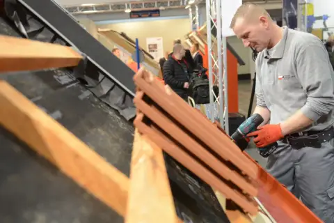 Dekarz w czerwonych rękawicach używa wkrętarki do montażu dachówek na konstrukcji dachu.