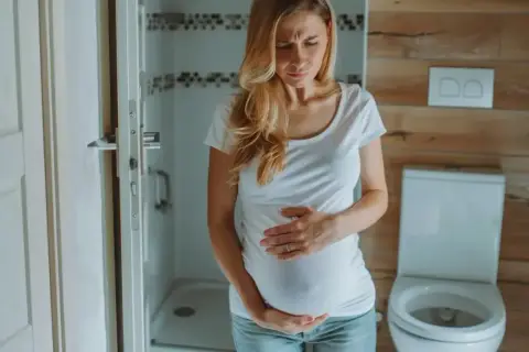 Kobieta w ciąży z wyrazem bólu na twarzy trzyma się za brzuch. W tle łazienka z toaletą. Ból podbrzusza w ciąży.