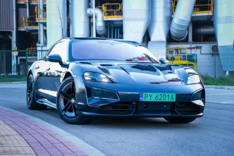 Elegancki Porsche Taycan w ciemnym kolorze, zaparkowany przed industrialnym budynkiem z rurami.