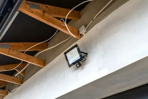 Nowoczesna elektryka w garażu: jasny reflektor LED z czujnikiem ruchu zamontowany na betonowej belce, otoczony drewnianą konstrukcją dachu i białymi przewodami.