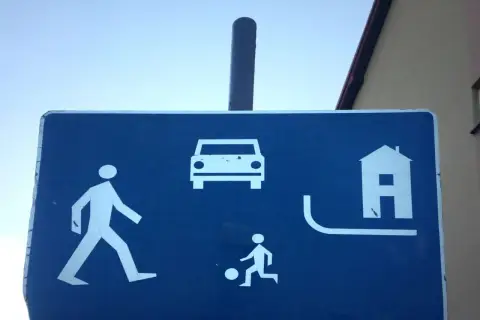 Znak informuje, że w strefie ruchu pieszy ma pierwszeństwo. Widać pieszego, dziecko grające w piłkę, samochód i dom.
