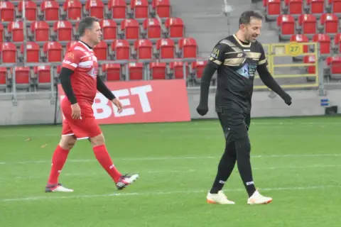 Dwóch piłkarzy, jeden w czerwonej koszulce z numerem 9, drugi w czarnej, idzie po boisku.
