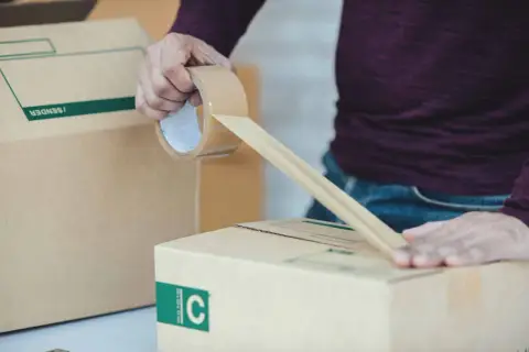 Osoba zakleja taśmą kartonowe pudło. To część procesu przygotowania przesyłki, który może być związany z odprawą celną.