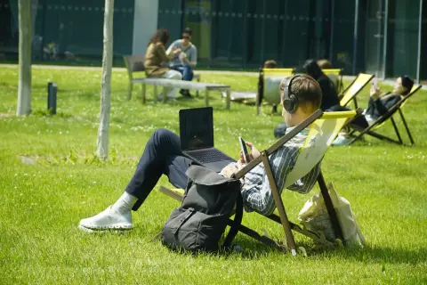 Mann mit Kopfhörern und Laptop im Freien, genießt sein Fernstudium im Ausland auf einer Liege im Sonnenschein.