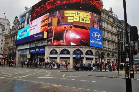 Tętniący życiem Piccadilly Circus w Londynie z neonowymi reklamami, w tym Samsung, McDonald's i Hyundai. Tłum ludzi na chodniku.