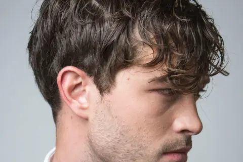 El pelo hombre, oscuro y ondulado, cae sobre su rostro. Se aprecian su oreja y parte de su barba incipiente.
