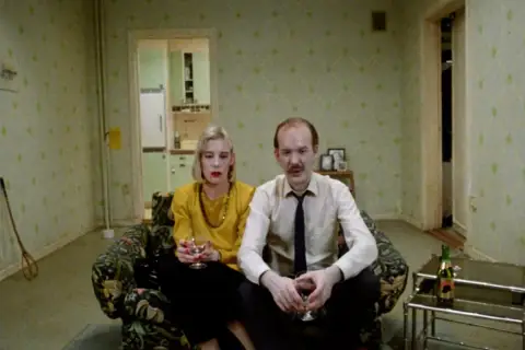 Ett par sitter i en soffa, en kvinna med gul tröja och en man i skjorta och slips. Kanske en scen från en Roy Andersson reklamfilm.