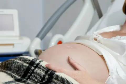 Kobieta w szpitalnym łóżku, z ręką na brzuchu, czeka na poród. Obok monitor KTG, który pomaga ocenić stan dziecka i może sugerować, jak przyspieszyć poród.