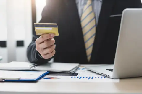 Biznesmen trzyma kartę kredytową, planując wykorzystać kredyt odnawialny do rozwoju firmy. Na biurku laptop i wykresy.