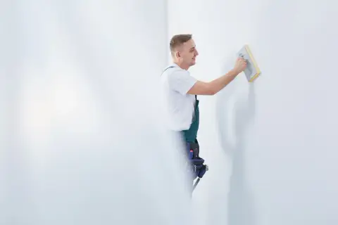 Fachowiec w zielonym ogrodniczkach wykonuje szlifowanie gładzi na białej ścianie, przygotowując ją do malowania.