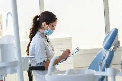 Studentka stomatologii w masce i okularach notuje coś na kartce. W tle widać fotel dentystyczny.