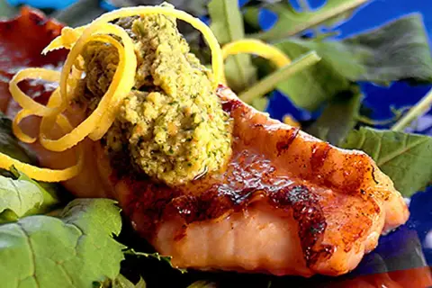 Grillad havsabborre med en grön topping och citronskal, serverad på en bädd av gröna blad.