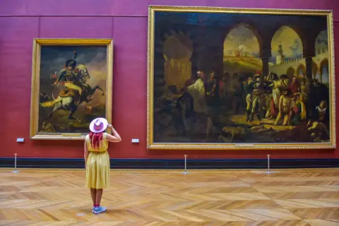 Kobieta w żółtej sukience i różowym kapeluszu podziwia obrazy w Luwrze. Co to za muzeum? To słynny paryski Luwr!