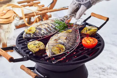 Deux poissons entiers grillent sur un barbecue, accompagnés d'artichauts et de tomates. Un gant tricoté ajuste la grille. Un vrai barbecue hivernal !