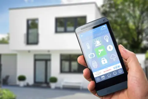 Dłoń trzyma smartfon z aplikacją do **instalacji inteligentnego domu**. Na ekranie widoczne ikony sterowania oświetleniem, ogrzewaniem, kamerami i innymi funkcjami.