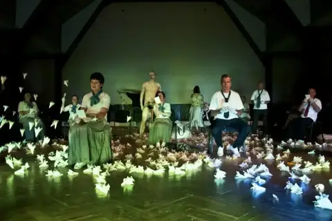 Scena teatralna pełna papierowych ptaków, ludzie w strojach z epoki i współczesnych ubraniach, posąg. To wszystko tworzy atmosferę teatru absurdu.