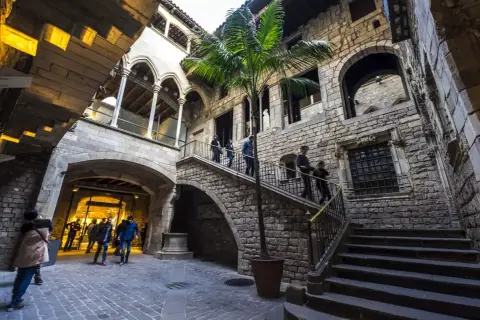 Un patio interior de piedra en el Born, con una palmera y escaleras que suben. Gente pasea por el lugar.