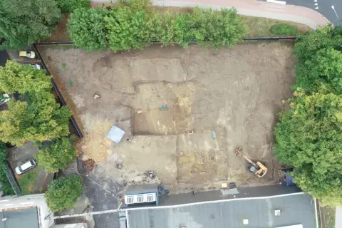 Wykop archeologiczny na placu budowy, gdzie prowadzone są badania przed pozwoleniem na budowę. Widoczny koparka i ślady dawnej zabudowy.