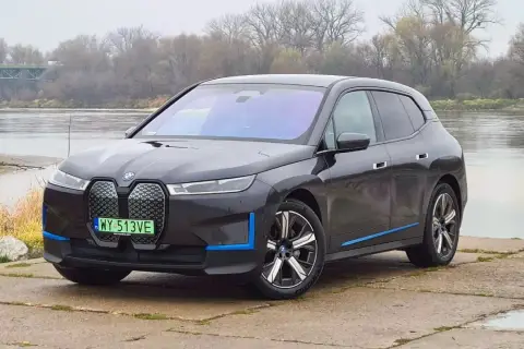 Ciemnoszary BMW iX z niebieskimi akcentami stoi nad rzeką, z mostem w tle.