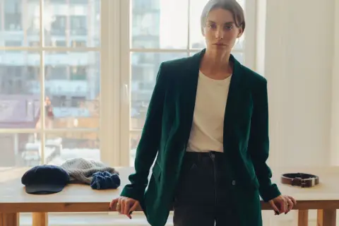 Elegancka kobieta w ciemnozielonej marynarce z aksamitu, stojąca przy drewnianym stole. Na stole czapka, szalik i pasek.
