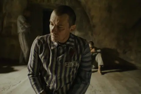 Film o Maksymilianie Kolbe: więźniowie w pasiakach, jeden z nich z numerem 16670, w ponurym wnętrzu.