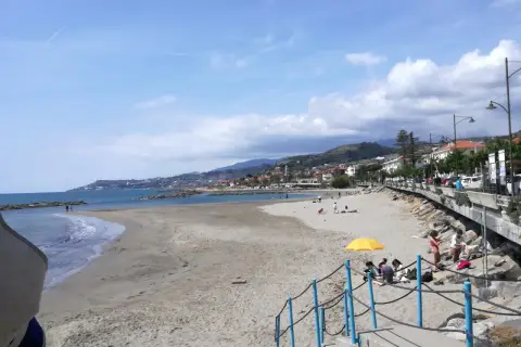 Plage de sable à Santo Stefano al Mare, avec des gens profitant du soleil et de la mer. La ville s'étend le long de la côte, bordée de collines verdoyantes.