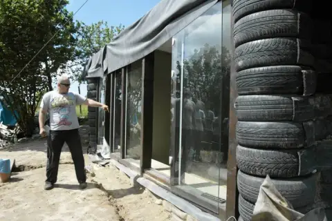 Budowa **earthship** z opon. Mężczyzna wskazuje na szklaną ścianę domu z opon.