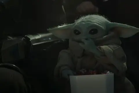 Dziecko z mandalorian film, w brązowym płaszczu, je słodycze z białego pudełka. Obok widać fragment zbroi Mandalorianina.