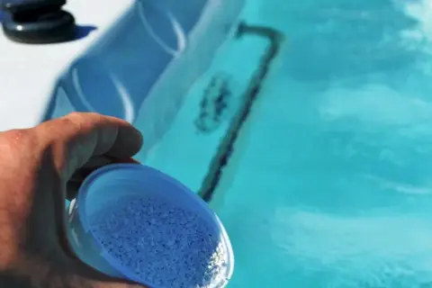 Dłoń wsypuje granulki bromu do błękitnej wody jacuzzi, dbając o czystość.