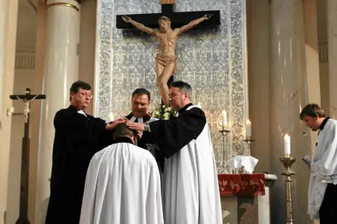 Trzech duchownych w szatach błogosławi klęczącego mężczyznę. Ceremonia w kościele protestanckim.