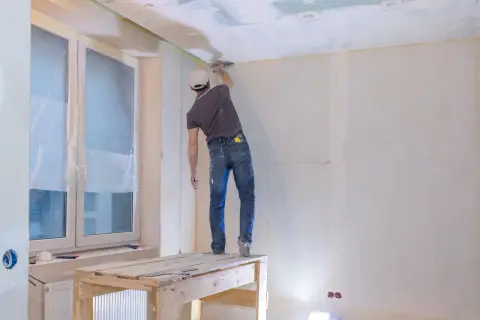 Pracownik na rusztowaniu wykonuje szpachlowanie łączeń płyt gipsowych na suficie.