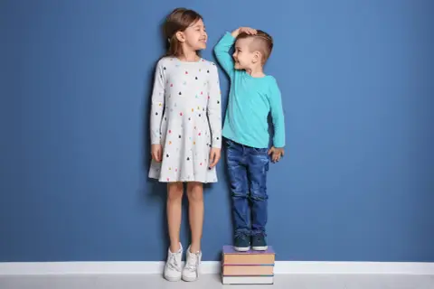 Dzieci mierzą wzrost. Chłopiec stoi na książkach, by dorównać siostrze. Wiedza to klucz, jak pobudzić wzrost dziecka.