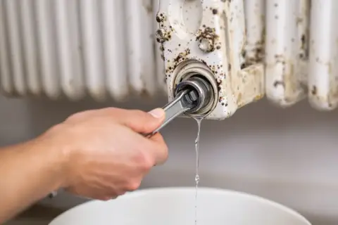 Ręka z kluczem odkręca zawór grzejnika, z którego kapie woda. Dowiedz się, jak wymienić zawór w grzejniku bez spuszczania wody.