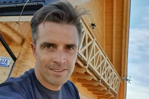 Staszek Karpiel-Bułecka pozuje na tle drewnianego domu z balustradą.