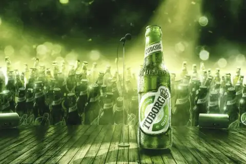 Zielona butelka piwa Tuborg Green na scenie, otoczona tłumem innych butelek. W tle mikrofon.