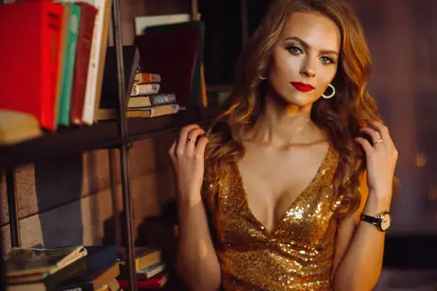 Elegancka kobieta w złotej sukience, z wyrazistym makijażem i czerwoną szminką, pozuje na tle regału z książkami.