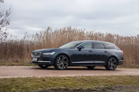 Ciemnogranatowe Volvo v90 na tle wysokiej, suchej trawy i pochmurnego nieba.