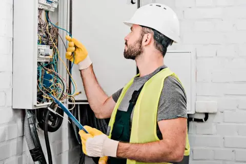 Elektryk w kasku i kamizelce odblaskowej pracuje przy skrzynce z przewodami. Zastanawia się, ile za punkt elektryczny.