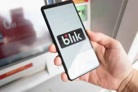 Dłoń trzyma smartfon z logo Blik przed bankomatem. Uważaj na oszustów, którzy mogą próbować wyłudzić dane.