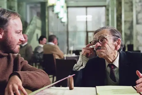 Dwóch mężczyzn rozmawia przy stole. Jeden z nich, starszy, w okularach, gestykuluje. Scena z jednego z filmów Krzysztofa Krauzego.