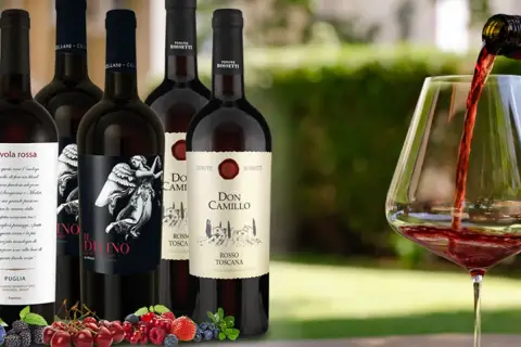 Flaschen mit teurem italienischem Rotwein, darunter "Il Divino" und "Don Camillo", werden vor einem Glas Rotwein präsentiert, in das gerade eingeschenkt wird. Frische Beeren und Kirschen liegen im Vordergrund.