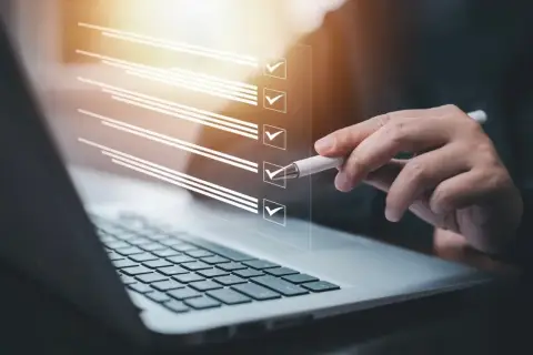 Dłoń z długopisem zaznacza pozycje na liście kontrolnej wyświetlanej nad klawiaturą laptopa.
