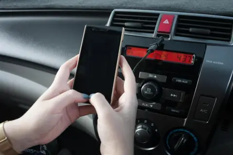 Ręce trzymają telefon, który jest podłączony kablem do radia samochodowego. Prosty sposób, jak połączyć telefon z samochodem.