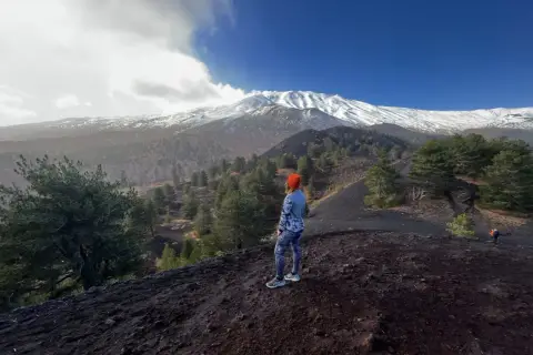 Kobieta w pomarańczowej czapce patrzy na ośnieżony wulkan Etna, otoczony lasem i czarną ziemią.