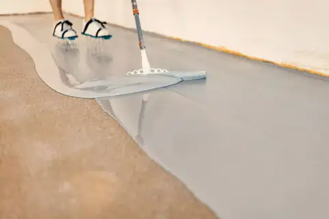Osoba w butach rozlewa szarą żywicę na betonową podłogę. Widać nierówności, które mogą być wadami podłogi z żywicy.