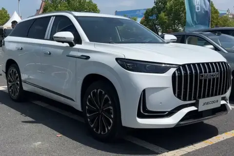 Biały SUV Jaecoo 9 z chromowanym grillem i czarnymi felgami stoi na parkingu.