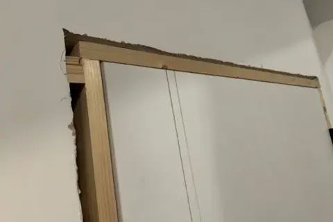 Widoczna szpara między ościeżnicą a ścianą, odsłaniająca drewnianą konstrukcję i fragment białych drzwi.